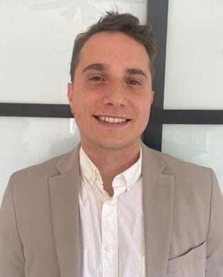 A person in a light-colored suit and white shirt stands in front of a window, smiling at the camera.